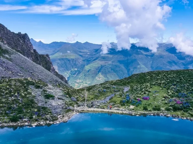 Laguna azul entre montañas rocosas y valles verdes de los Andes, con nubes sobre los picos