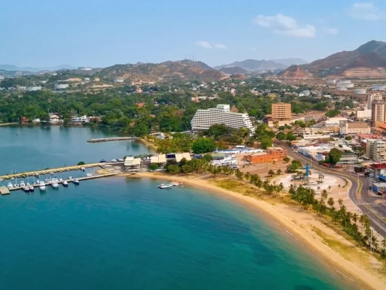 Vista aérea de una playa con muelle de lanchas, un hotel blanco escalonado y la ciudad entre colinas